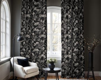 Op de afbeelding: Zwart-witte bloemen gordijnen met een schedelpatroon. De gordijnen hangen in een woonkamer met een witte fauteuil, een zwarte lamp en een kleine tafel met een vaas met bloemen.
