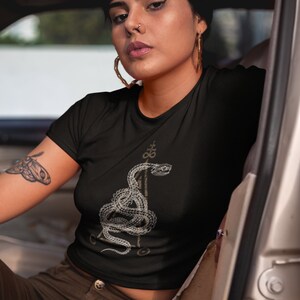 May include: A black cropped t-shirt with a white graphic of a snake wrapped around a triangle. The snake has a stylized design and is surrounded by other symbols.