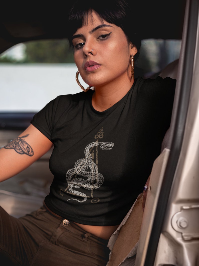 May include: A black cropped t-shirt with a white graphic of a snake wrapped around a triangle. The snake has a stylized design and is surrounded by other symbols.