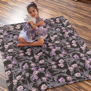 Child sitting on a wooden floor holding stuffed animal sitting on the pastel goth blanket that has pink, purple, and blue skull flowers.