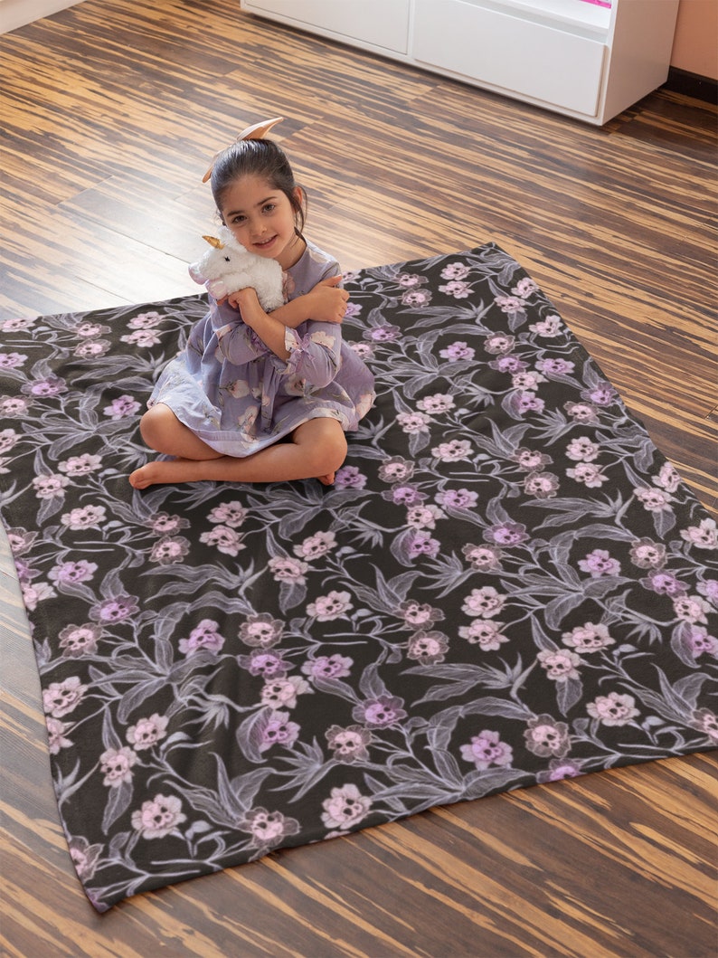 Child sitting on a wooden floor holding stuffed animal sitting on the pastel goth blanket that has pink, purple, and blue skull flowers.