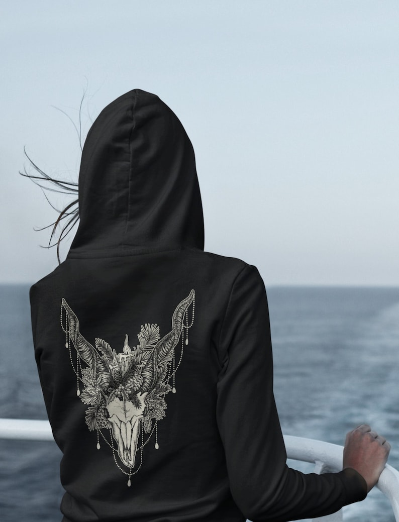 May include: Black hoodie with a detailed, cream-colored illustration on the back. The design features a skull with large horns, adorned with floral and beaded elements. The person is standing near the water.