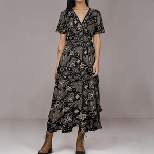 Puede incluir: Vestido cruzado negro con cuello en V y mangas cortas. El vestido presenta un patrón repetido de polillas, lunas y soles en color beige. El vestido se lleva con botas marrones.