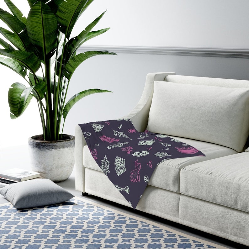 May include: A dark purple throw blanket with a white and pink pattern of various symbols and designs. The blanket is draped over a white sofa.