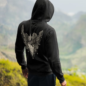 May include: Black hooded sweatshirt with a detailed, cream-colored graphic on the back featuring a skull, horns, and floral elements. The design is intricate and appears to be a pen and ink style illustration. The sweatshirt is worn by a person facing away from the camera.