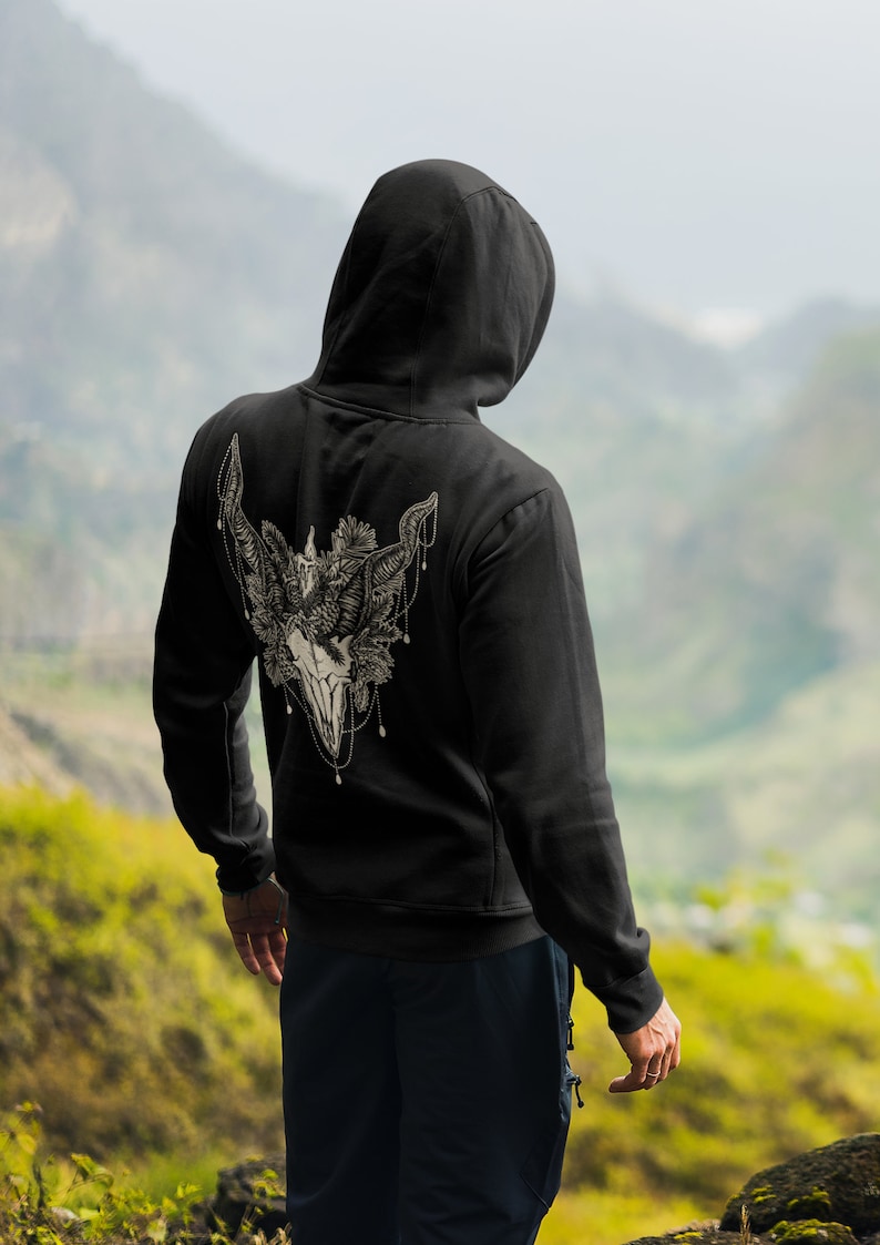 May include: Black hooded sweatshirt with a detailed, cream-colored graphic on the back featuring a skull, horns, and floral elements. The design is intricate and appears to be a pen and ink style illustration. The sweatshirt is worn by a person facing away from the camera.