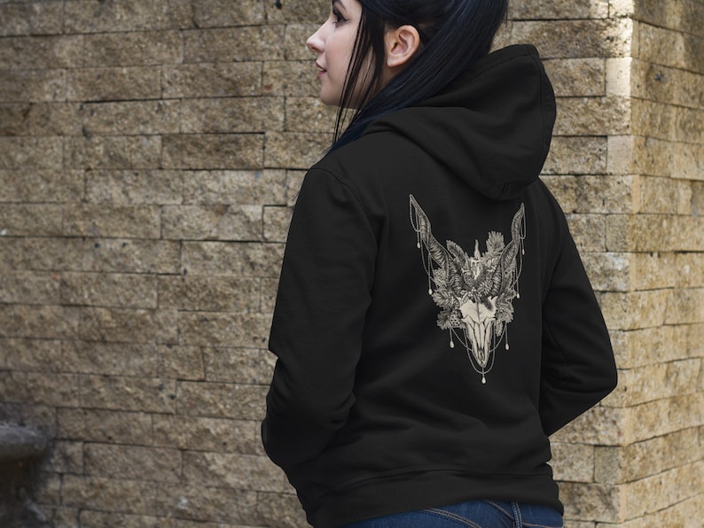 May include: Black hoodie with a detailed white graphic on the back featuring a skull, feathers, and floral elements. The design is intricate and centered on the upper back of the hoodie. The hoodie has a hood and a simple, classic design.