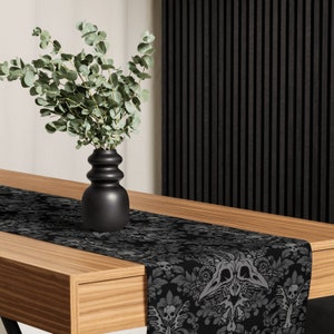 Puede incluir: Un camino de mesa negro y gris con un patrón de calavera y huesos. Un jarrón con hojas verdes se encuentra sobre una mesa de madera.