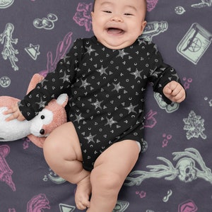 May include: A baby wearing a black long-sleeved onesie with a white star and crescent moon pattern. The onesie is on a dark purple background with a variety of white and pink illustrations.