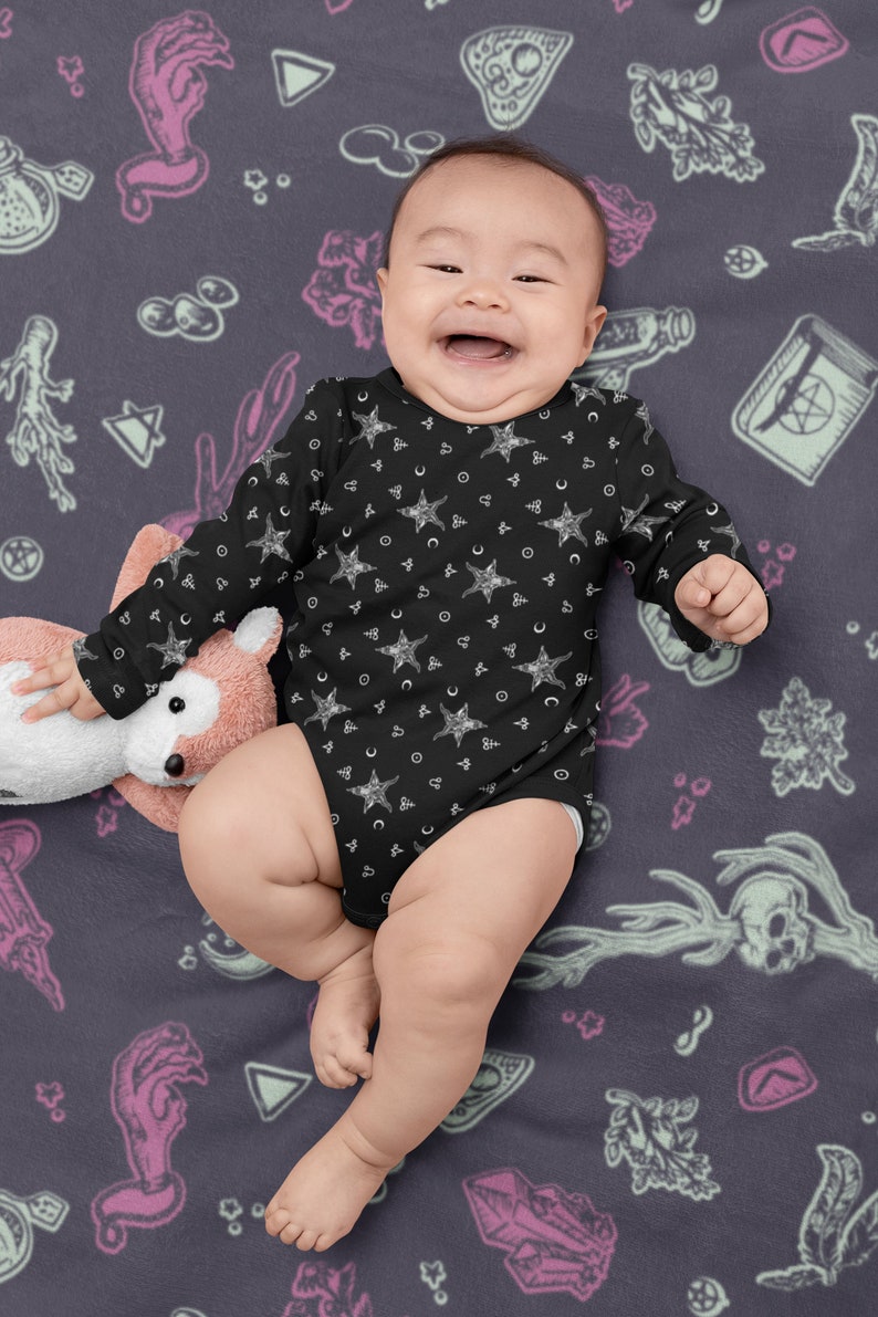 May include: A baby wearing a black long-sleeved onesie with a white star and crescent moon pattern. The onesie is on a dark purple background with a variety of white and pink illustrations.