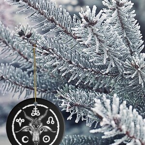 May include: A black ceramic Christmas ornament with a white design of a goat's head with horns, a pentagram, and other symbols. The ornament is hanging from a gold string on a snowy pine tree branch.