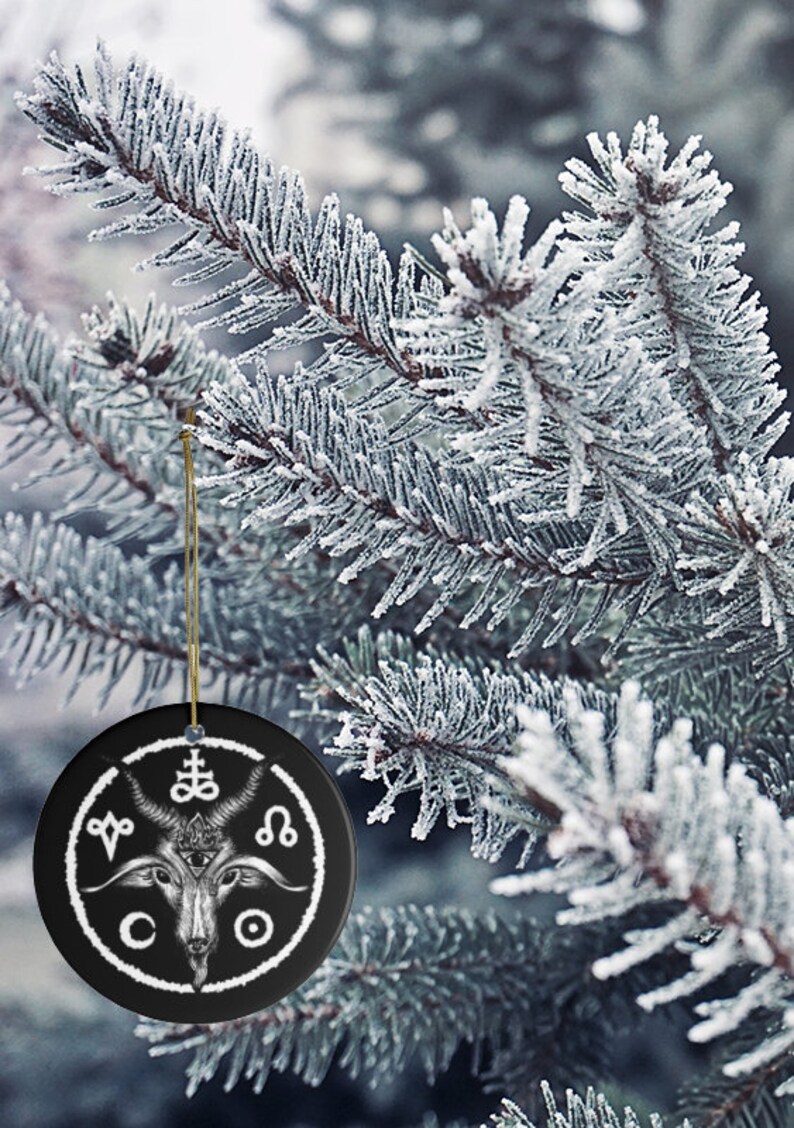 May include: A black ceramic Christmas ornament with a white design of a goat's head with horns, a pentagram, and other symbols. The ornament is hanging from a gold string on a snowy pine tree branch.