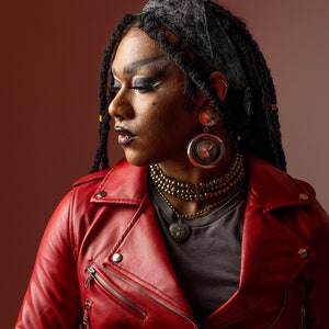 May include: A person with dreadlocks wearing a red leather jacket, a black bandana, and silver earrings. They are wearing a gray t-shirt and a gold beaded necklace with a silver heart pendant.