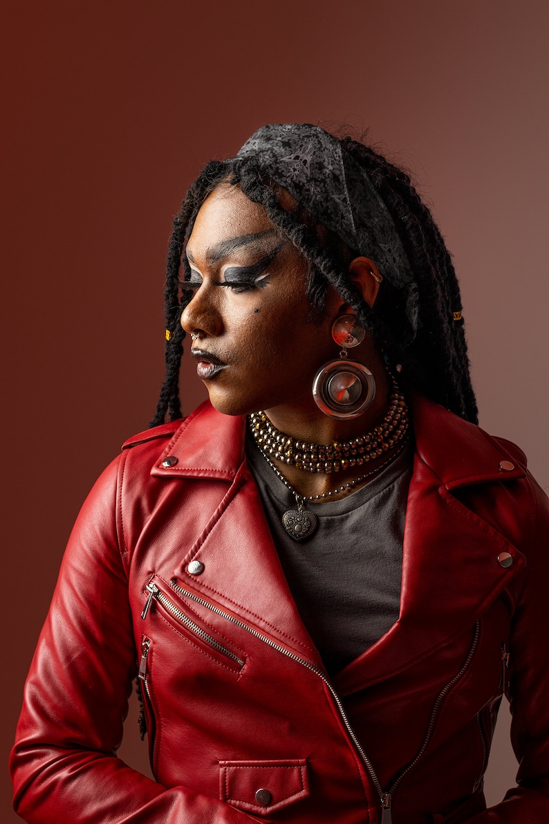 May include: A person with dreadlocks wearing a red leather jacket, a black bandana, and silver earrings. They are wearing a gray t-shirt and a gold beaded necklace with a silver heart pendant.