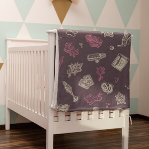 May include: A white cot with a grey and pink patterned blanket featuring illustrations of crystals, herbs, and tarot cards. The blanket is folded over the cot and has a white border.