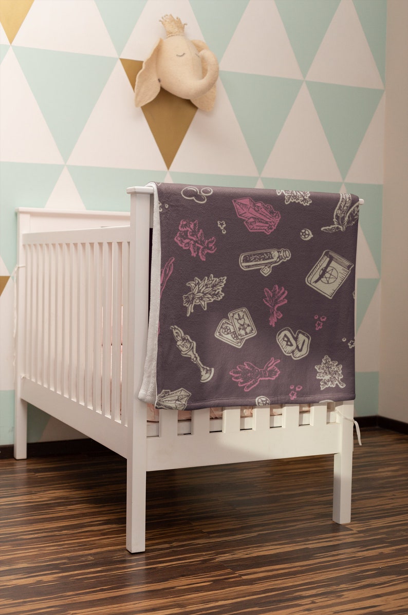 May include: A white cot with a grey and pink patterned blanket featuring illustrations of crystals, herbs, and tarot cards. The blanket is folded over the cot and has a white border.