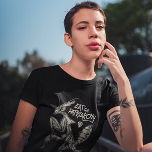 Może przedstawiać: Czarny T-shirt z białym nadrukiem przedstawiającym modliszkę z tekstem "Eat the Patriarchy" i symbolem żeńskim.