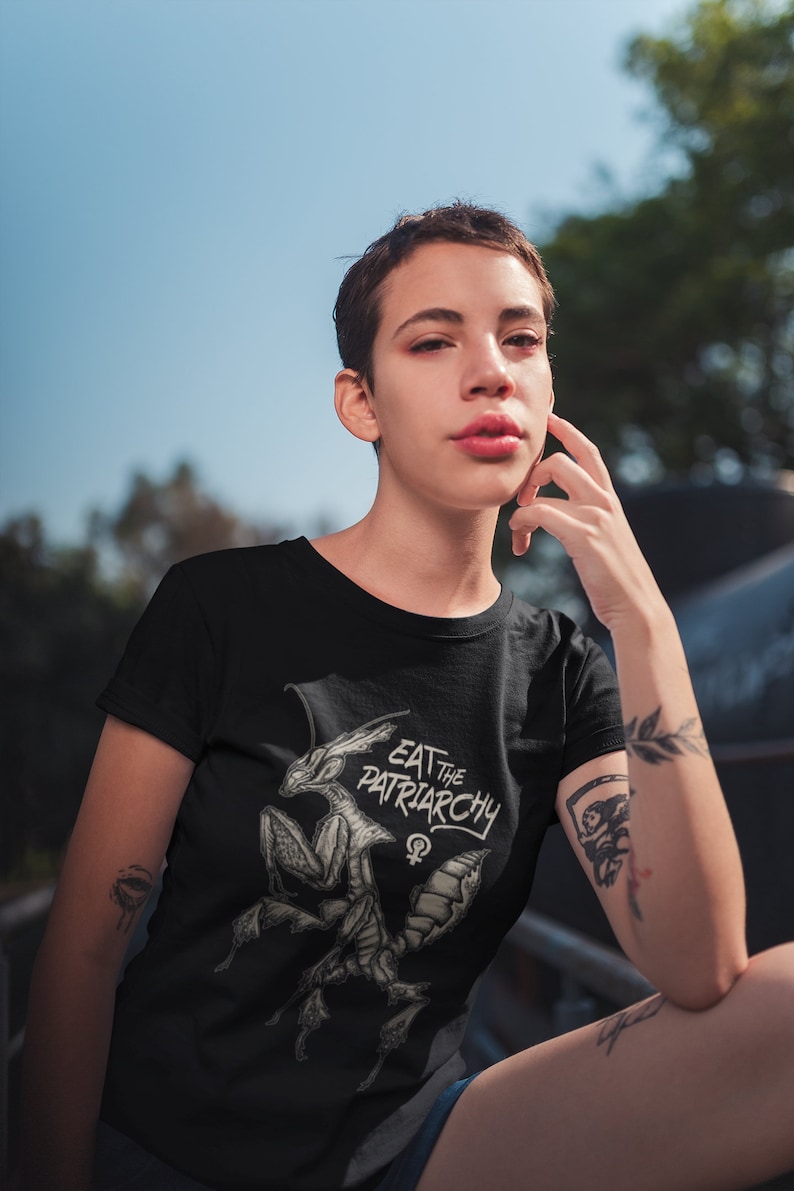 Może przedstawiać: Czarny T-shirt z białym nadrukiem przedstawiającym modliszkę z tekstem "Eat the Patriarchy" i symbolem żeńskim.
