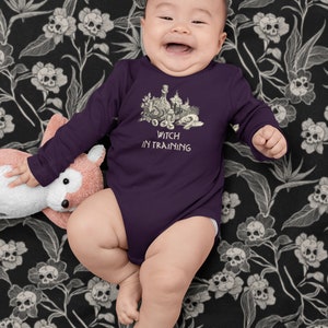 May include: A purple long-sleeved baby onesie with a black and white illustration of a witch's cauldron and the text "Witch in Training". The baby is wearing the onesie and is sitting on a black and white floral blanket.