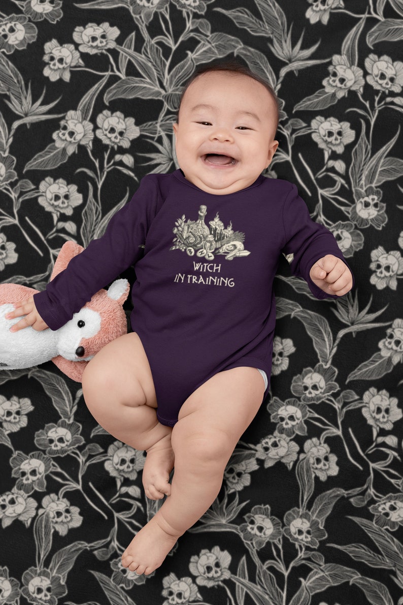 May include: A purple long-sleeved baby onesie with a black and white illustration of a witch's cauldron and the text "Witch in Training". The baby is wearing the onesie and is sitting on a black and white floral blanket.