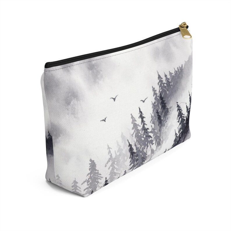 May include: A white zippered pouch with a black and gray watercolor design of a forest with trees and birds. The pouch is made of canvas and has a zipper closure.