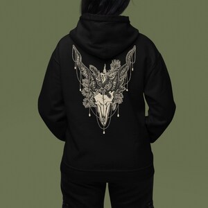 May include: Black hoodie featuring a detailed cream-colored illustration of a skull with large horns, adorned with floral and beaded accents. The design is centered on the back of the hoodie, offering a gothic and nature-inspired aesthetic.