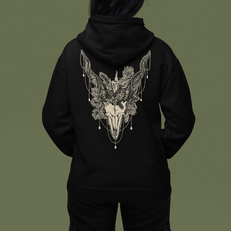 May include: Black hoodie featuring a detailed cream-colored illustration of a skull with large horns, adorned with floral and beaded accents. The design is centered on the back of the hoodie, offering a gothic and nature-inspired aesthetic.