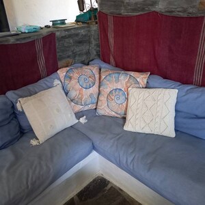 May include: A blue sectional sofa with four pillows. Two of the pillows have a spiral shell design, one is white with tassels, and one is white with a diamond pattern. The sofa is against a stone wall with a red and white striped fabric hanging behind it.
