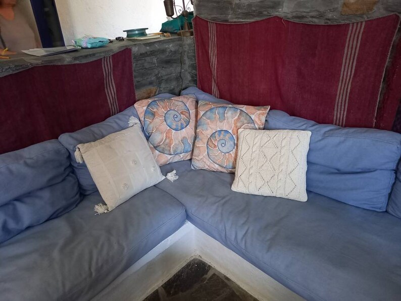 May include: A blue sectional sofa with four pillows. Two of the pillows have a spiral shell design, one is white with tassels, and one is white with a diamond pattern. The sofa is against a stone wall with a red and white striped fabric hanging behind it.