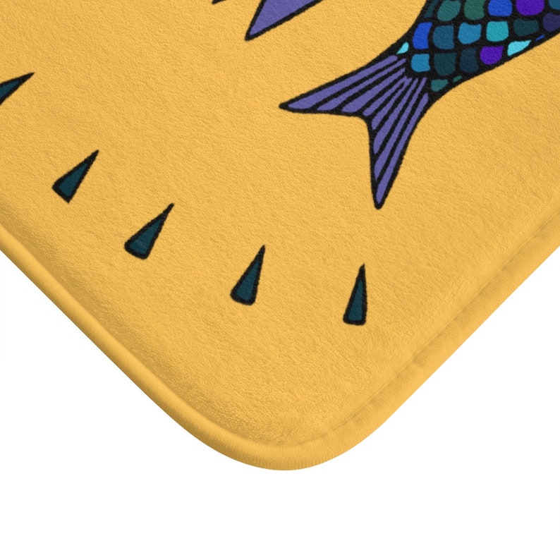 Puede incluir: Una alfombra de ba&ntilde;o amarilla con un dise&ntilde;o de pez azul y morado. El pez tiene escamas y una cola morada.