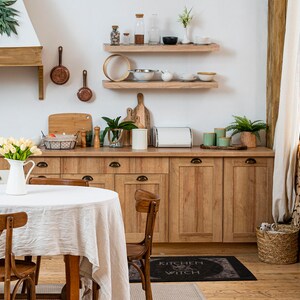 May include: A white kitchen table with a white tablecloth and four chairs. The table is set with a jug of flowers and a rug with the text "Kitchen Witch" on it.