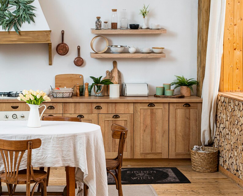 May include: A white kitchen table with a white tablecloth and four chairs. The table is set with a jug of flowers and a rug with the text "Kitchen Witch" on it.