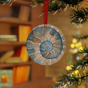 May include: A round, blue and brown watercolour ornament featuring a spiral ammonite fossil design. The ornament is hanging from a red ribbon on a Christmas tree.