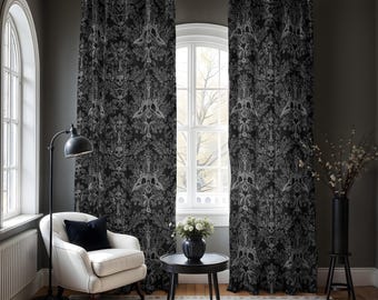 Op de afbeelding: Een paar zwarte gordijnen met een wit schedel- en bloemenpatroon hangen in een woonkamer met een witte fauteuil, een zwarte bijzettafel en een vaas met witte bloemen.