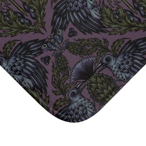 May include: A rectangular mat featuring a repeating pattern on a dark purple background. The design includes illustrations of black crows with blue-tinted feathers, green thistle plants, small dark berries, and moths with skull-like markings.