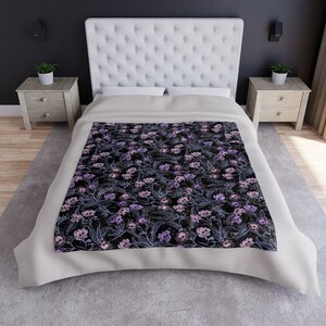 Laid out pastel blanket with purple, pink and blue skull flowers made of crushed velvet. Blanket is laid on bed with grey rug beneath the bed.