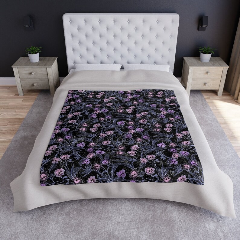 Laid out pastel blanket with purple, pink and blue skull flowers made of crushed velvet. Blanket is laid on bed with grey rug beneath the bed.