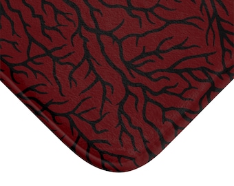 May include: A burgundy bath mat with a black, branching pattern. The mat has a textured surface and a rounded corner, suggesting a comfortable and stylish design for a bathroom or similar space.