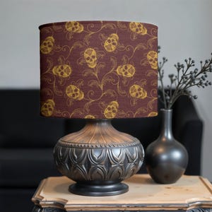 Può includere: Un paralume marrone scuro con un motivo di teschio dorato e fiori. Il paralume è su una base di lampada nera con un design ornato.