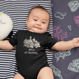 May include: A baby wearing a black bodysuit with a white graphic of a witch's cauldron and the text "Witch in Training". The baby is lying on a striped blanket with a colourful pillow.