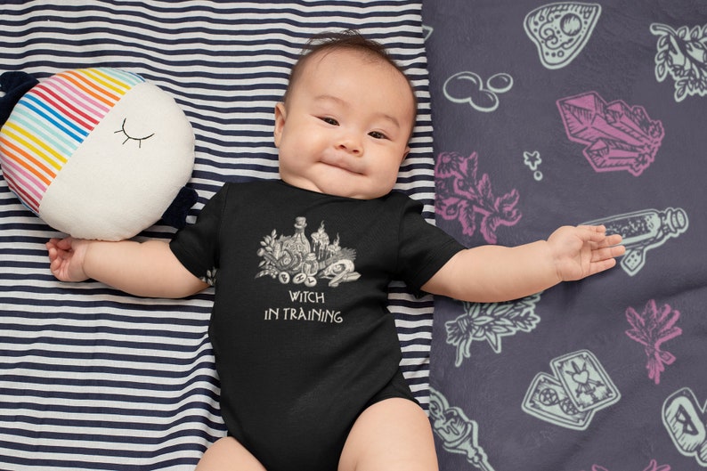 May include: A baby wearing a black bodysuit with a white graphic of a witch's cauldron and the text "Witch in Training". The baby is lying on a striped blanket with a colourful pillow.