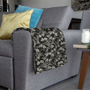 May include: A gray sofa with two throw pillows, one yellow and one blue. A black and white throw blanket with a skull and flower pattern is draped over the arm of the sofa.