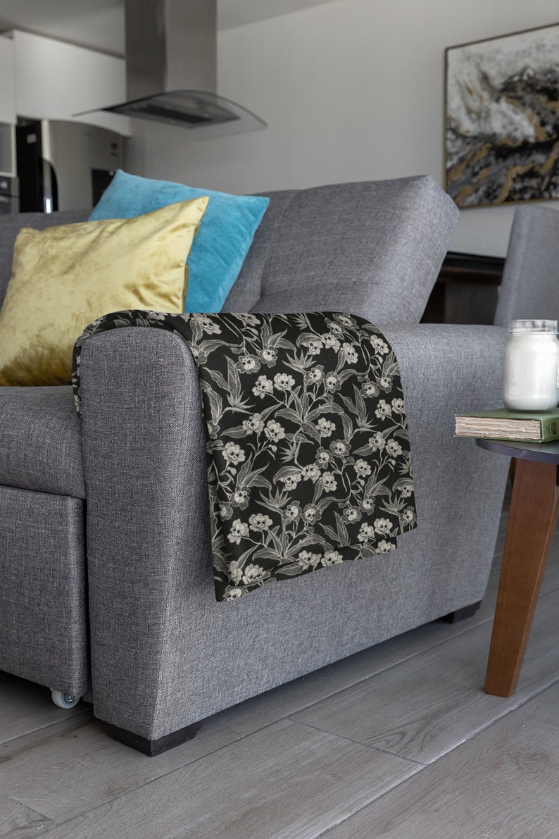 May include: A gray sofa with two throw pillows, one yellow and one blue. A black and white throw blanket with a skull and flower pattern is draped over the arm of the sofa.