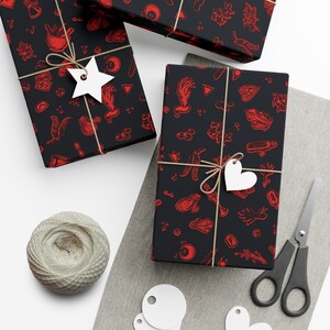 May include: Three black gift boxes wrapped in red and black patterned paper with a spooky theme. The paper features illustrations of hands, crystals, and other occult symbols. The boxes are tied with twine and decorated with white star and heart-shaped gift tags.