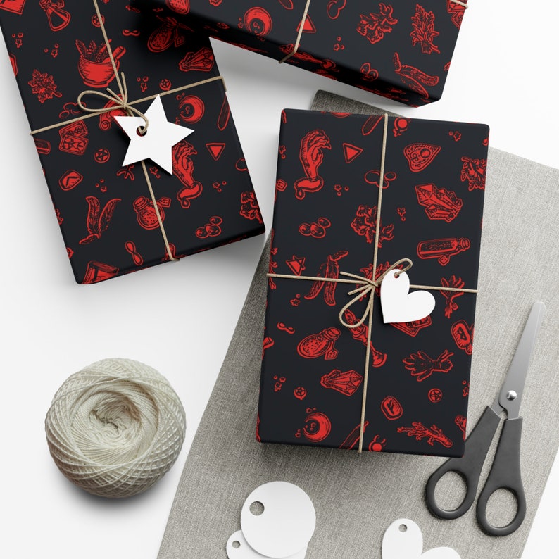May include: Three black gift boxes wrapped in red and black patterned paper with a spooky theme. The paper features illustrations of hands, crystals, and other occult symbols. The boxes are tied with twine and decorated with white star and heart-shaped gift tags.
