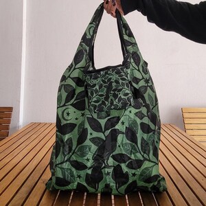 May include: Green and black reusable shopping bag with a leaf and raven pattern. The bag features a pocket and black handles. The design includes stars and a crescent moon. The bag is made of a lightweight material.