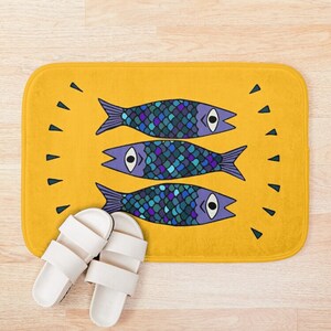 Puede incluir: Una alfombra de ba&ntilde;o amarilla con un dise&ntilde;o de tres peces azules y morados con escamas y ojos. Los peces est&aacute;n dispuestos en fila, con las colas apuntando hacia la derecha y las cabezas hacia la izquierda.
