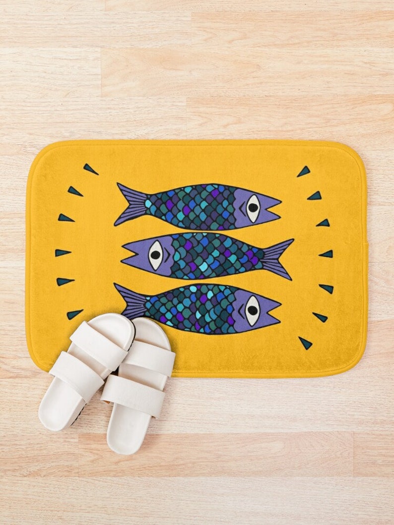 Puede incluir: Una alfombra de ba&ntilde;o amarilla con un dise&ntilde;o de tres peces azules y morados con escamas y ojos. Los peces est&aacute;n dispuestos en fila, con las colas apuntando hacia la derecha y las cabezas hacia la izquierda.