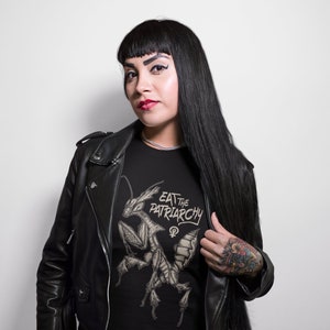 Może przedstawiać: Czarny T-shirt z grafiką modlącej się modliszki i tekstem "Eat the Patriarchy" w stylizowanym foncie. Koszulkę nosi osoba z długimi czarnymi włosami i czarną sk&oacute;rzaną kurtką.