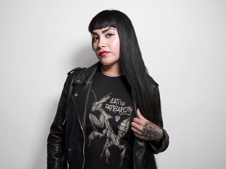 Może przedstawiać: Czarny T-shirt z grafiką modlącej się modliszki i tekstem "Eat the Patriarchy" w stylizowanym foncie. Koszulkę nosi osoba z długimi czarnymi włosami i czarną sk&oacute;rzaną kurtką.